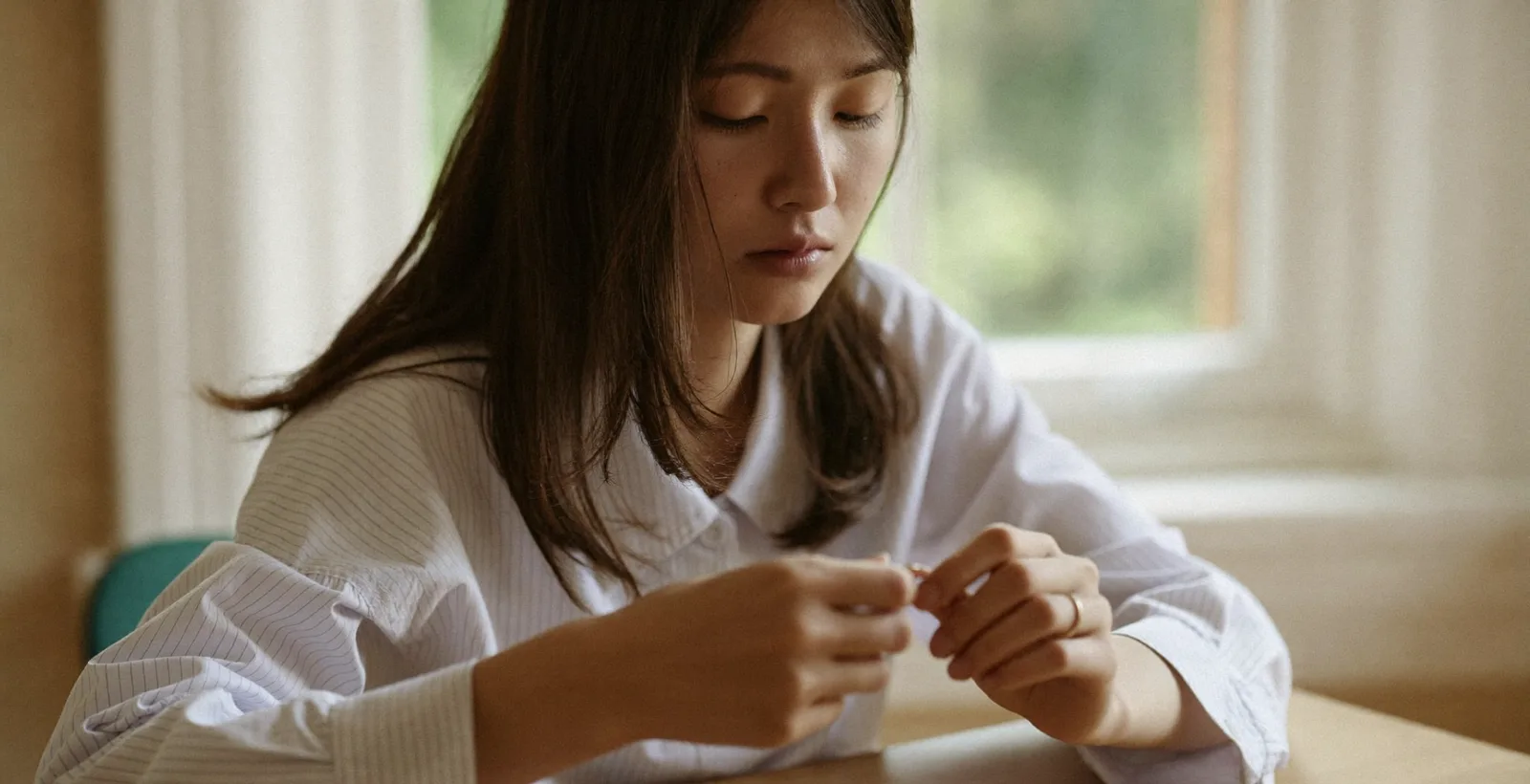 Thoughtful young woman by a window holding an engagement ring, reflecting quietly on commitment