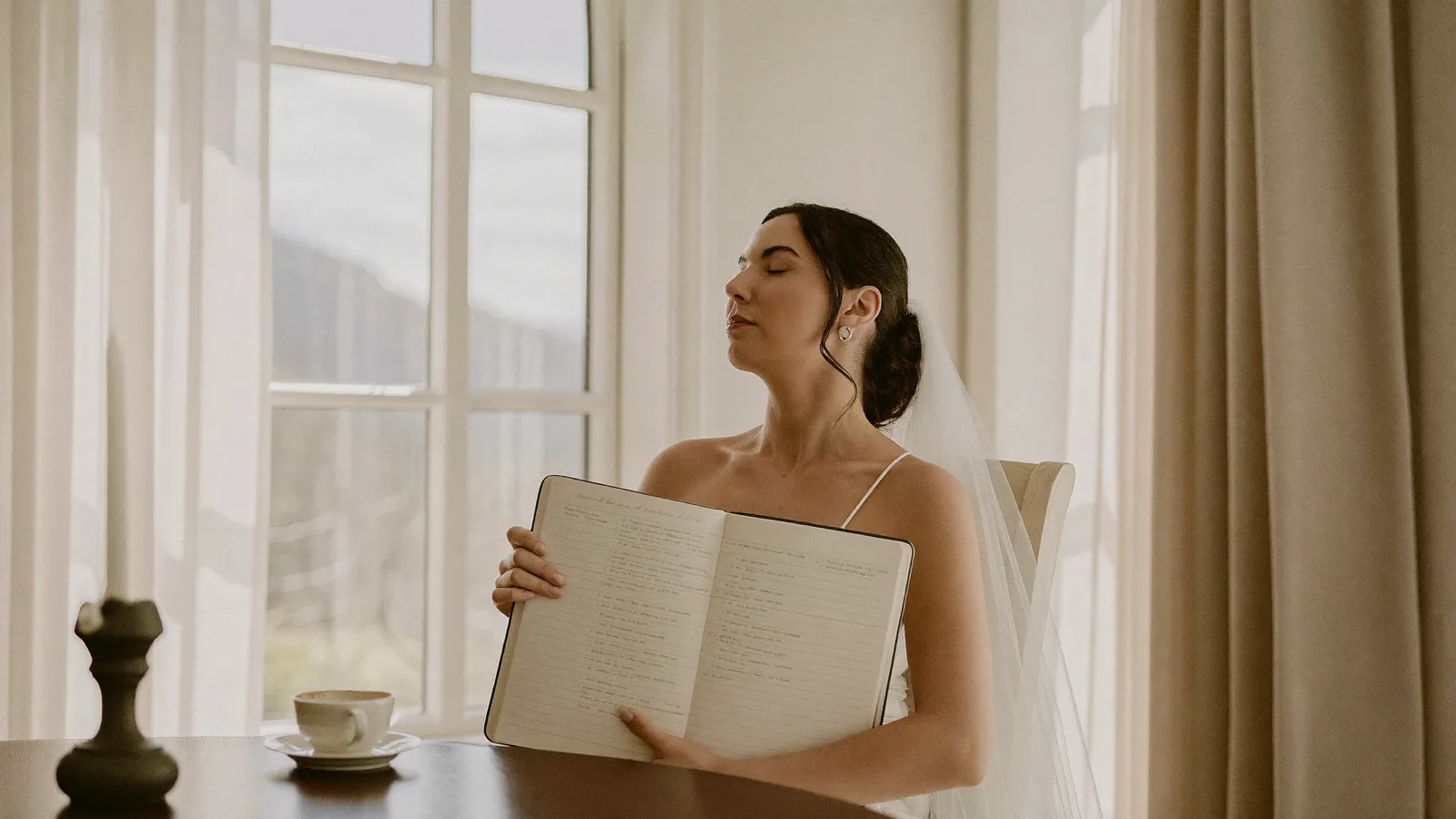 Bride-to-be taking a slow calming breath by a bright window, wedding checklist and tea on the table beside her